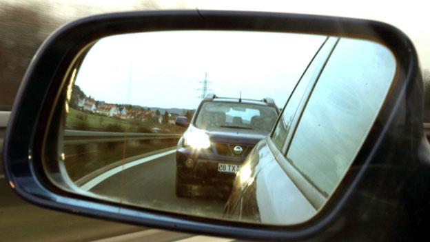 Im Außenspiegel eines Kraftfahrzeugs ist  ein dicht auffahrender Drängler zu sehen