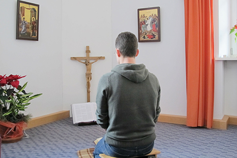 Junger Mann kniet in einem kargen Raum vor einem Kreuz