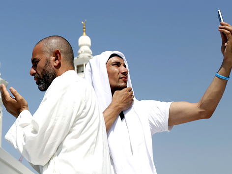 Zwei Männer, einer betend, einer ein Selfie schießend, beim Hadsch in Mekka