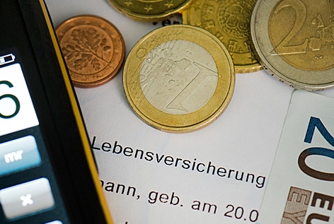 Auf einem Versicherungsschein für eine Lebensversicherung liegt ein Smartphone auf dem ein Taschenrechner angezeigt wird, sowie mehrere Euromünzen und ein Euroschein