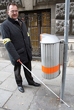 Ein Mann mit Blindenstock demonstriert die Hindernisse für Sehbeeinträchtigte im Straßenverkehr (in diesem Fall ein Mistkübel)