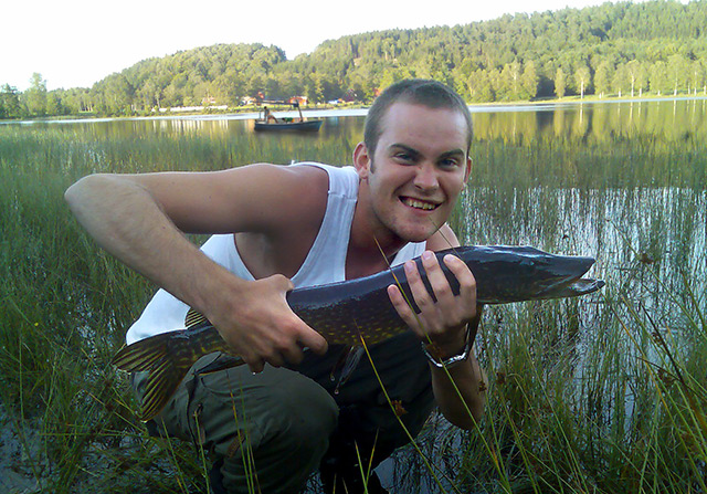 Junger Mann hält gefangenen Fisch in Händen, dahinter ein See