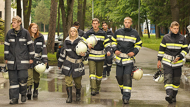 8 Junge Feuerwehrleute in Uniform und mit Helmen in der Hand kommen auf den Betrachter zu
