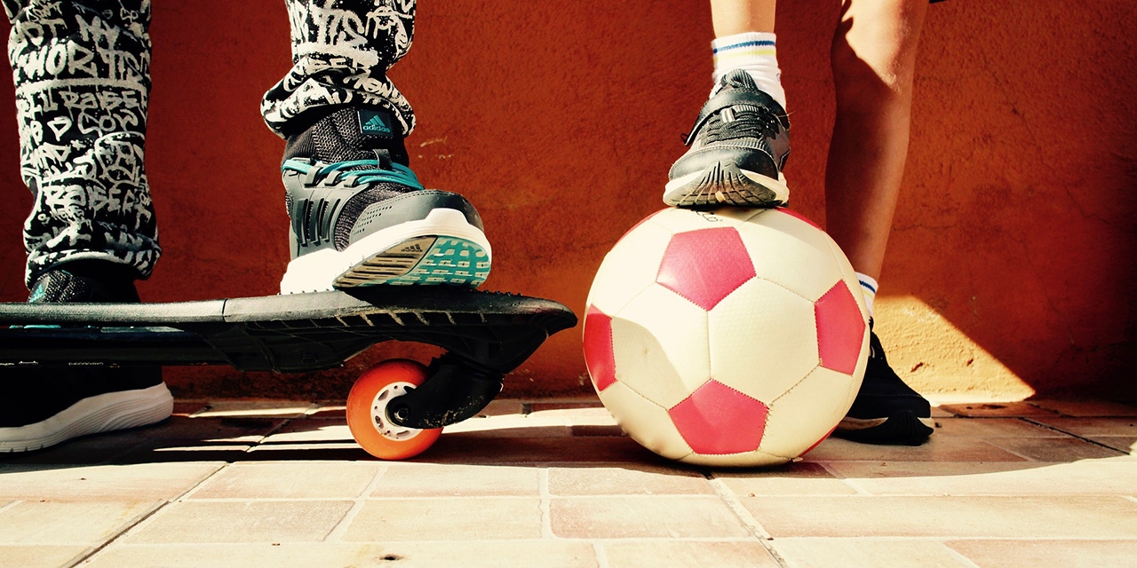 Die Füße von zwei Buben, einer mit Skateboard, der andere mit einem Fußball.