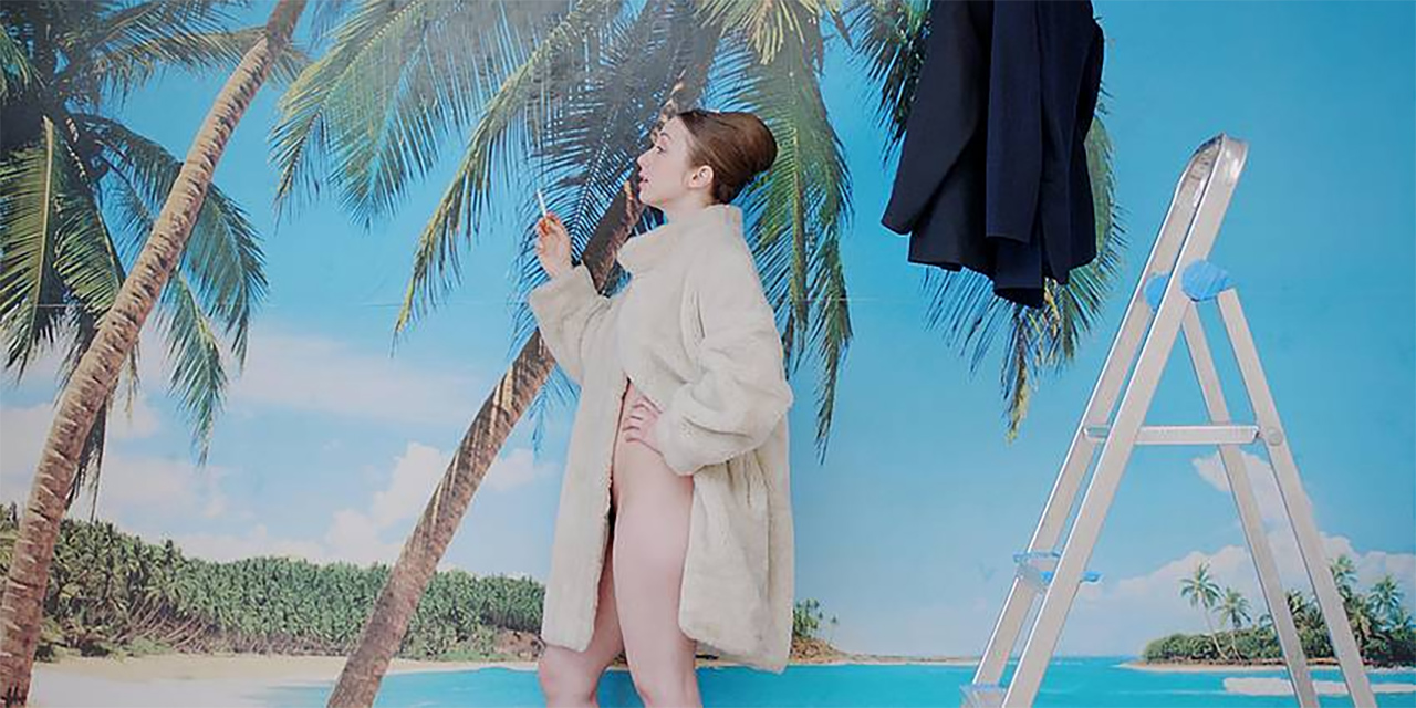 Frau mit nichts als einem Mantel bekleidet, vor einer Fototapete mit Palmen und Strand