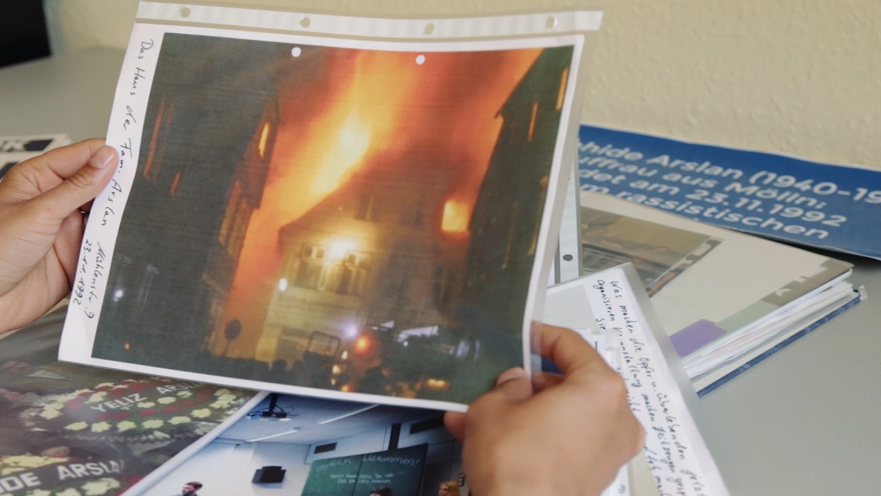 Hände halten eine Klarsichtfolie, in der das Bild eines brennendes Hauses ist. Das Bild zeigt den Brandanschlag von Mölln 1992.