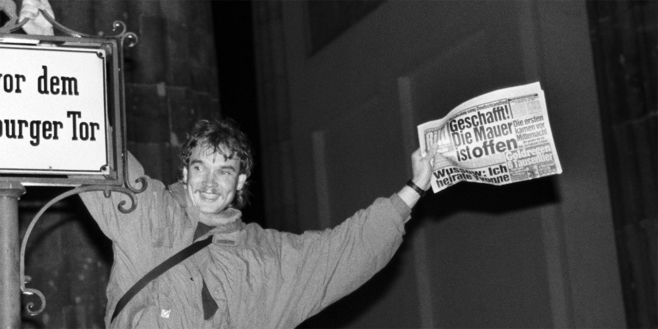 Ein Mann hält sich am 9.11.1989 in Berlin an dem Schild mit der Aufschrift "Platz vor dem Brandenburger Tor" fest und schwenkt eine BILD Zeitung mit dem Titel "Geschafft! Die Mauer ist offen".