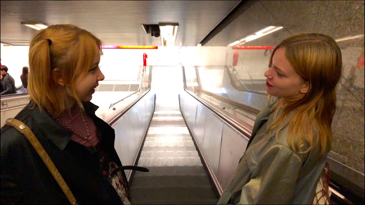 Zwei junge Frauen stehen auf einer Rollstreppe und lächeln einander an. Filmstill aus "Lololol".