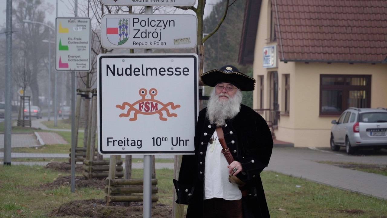 Ein Mann mit viel Bart und im Piratenkostüm steht neben einem Schild mit der Aufschrift "Nudelmesse 10 Uhr" an einem Ortsanfang. Filmszene aus "I, Pastafari".
