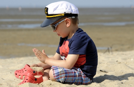 Ein Kind mit Kappe, Sonnenbrille und dunklem T-Shirt spielt am Strand