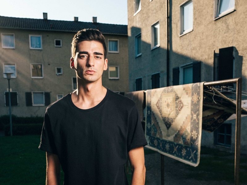 Der Autor Cihan Acar steht vor einer Wohnsiedlung, hinter ihm hängt ein Teppich auf einer Wäscheleine