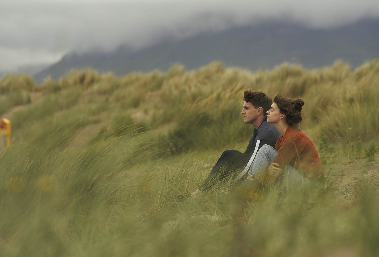 Filmszene aus der Serie "Normal People": eine junge Frau und ein junger Mann sitzen auf einem grünen Hügel