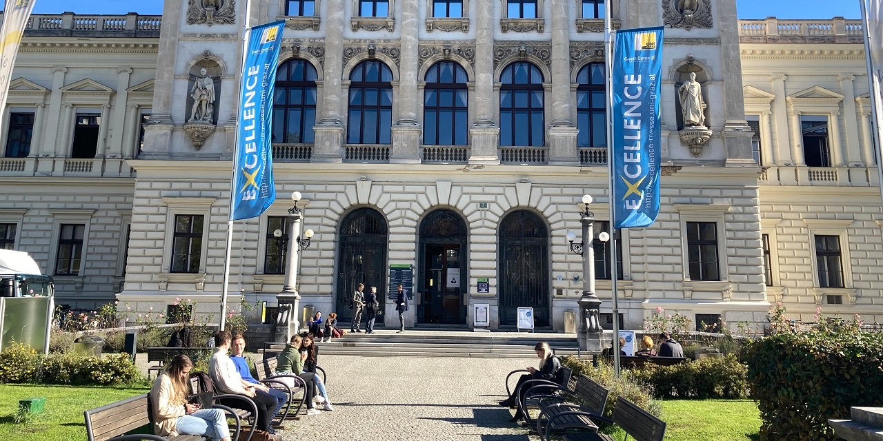 Das Hauptgebäude der Karl-Franzens-Universität in Graz, davor sitzen Studiererende auf den Bänken und unterhalten sich.