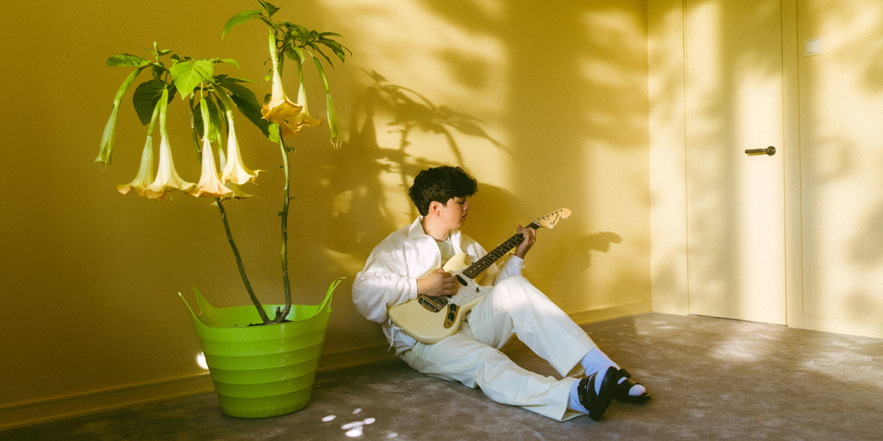 Boy Pablo sitzt neben einer blühenden Zimmerpflanze am Boden und spielt auf einer Gitarre
