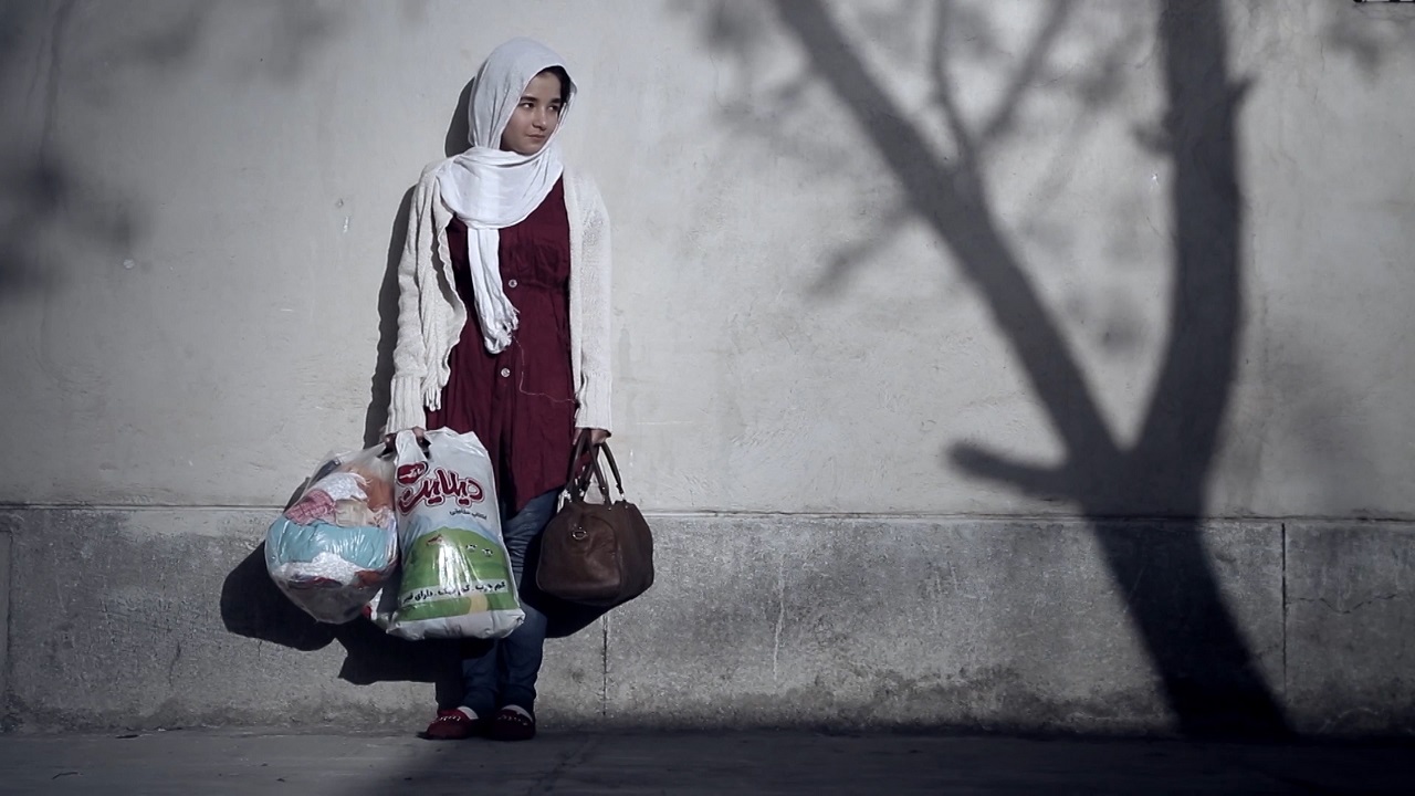 Ein iranisches Mädchen steht mit zwei Plastiksackerln mit ihrem Hab und Gut auf der Straße und lehnt an eine Hauswand.