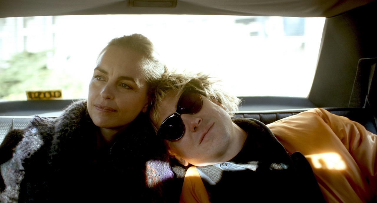 Lars Eidinger trägt eine blonde Perücke und dunkle Brillen und lehnt sich bei Nina Hoss an. Sie fahren im Taxi. Szene aus "Schwesterlein".