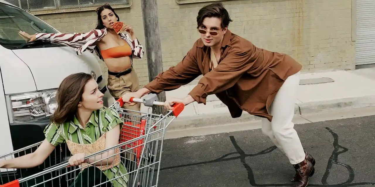 Naomi Higgins sitzt in einem Einkaufswagen, den Wil King schiebt und Olivia Junkeer isst eine Pizzaschnitte. Alle drei spielen in der Serie "Why Are You Like This".