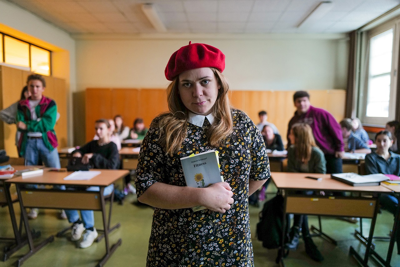 Stefanie Sargnagel steht mit einem Buch in der Hand in einer Schulklasse. Szene aus dem Film "Sargnagel".