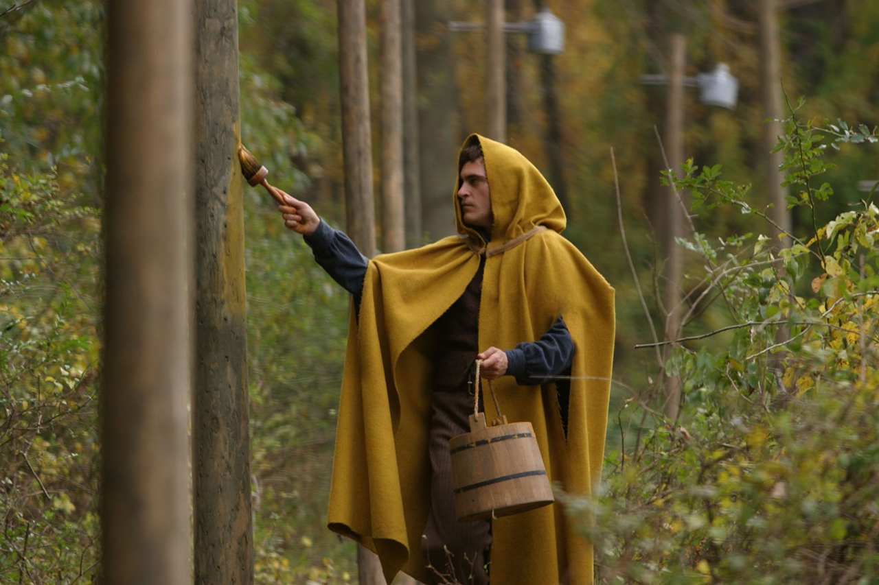 Ein junger Mann mit einem Kapuzenmantel steht im Wald und streicht mit einem Pinsel Farbe auf einen Baumstamm