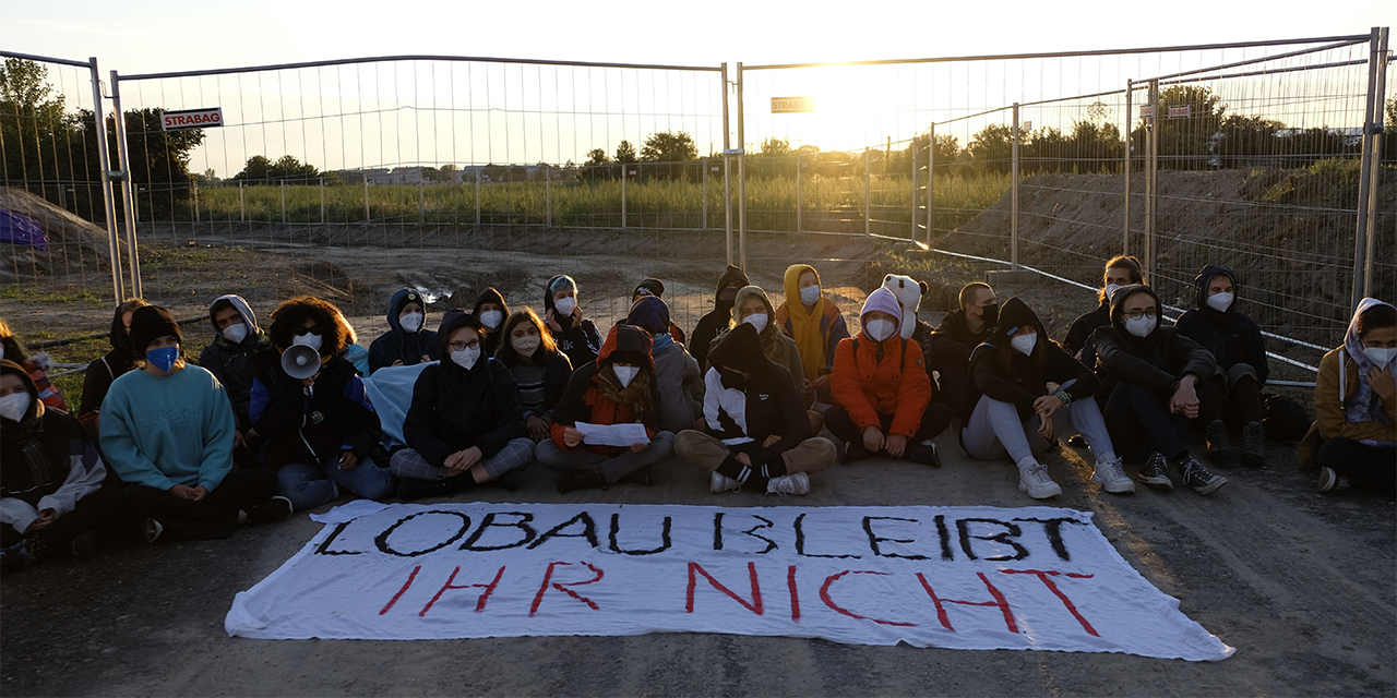 Eine Gruppe von Klimaaktivistinnen und Aktivisten sitzt vor einem Transparent mit der Aufschrift: "Lobau bleibt - ihr nicht"