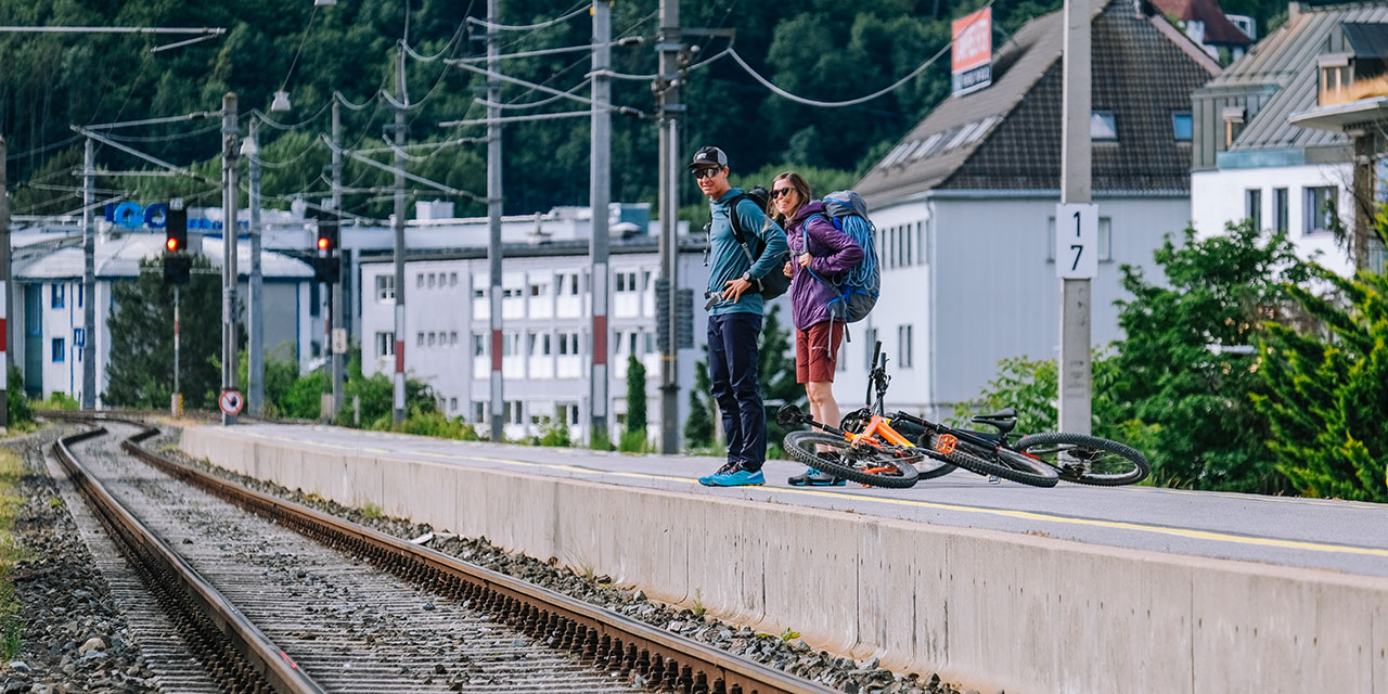 Zwei Bergsportler*innen warten am Bahnsteig