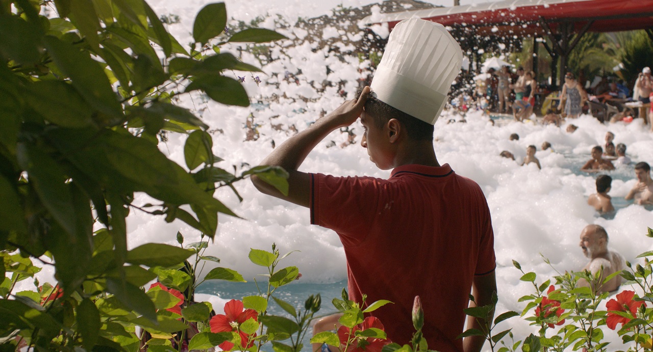 Ein 18-jähriger Kochlehrling steht mit Haube am Rand eines Pools, in dem Urlauber eine Schaumparty feiern. Szene aus der Doku "All-In".