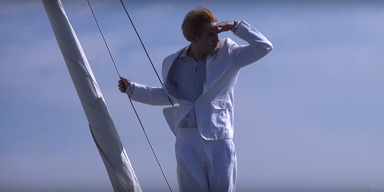 Ein junger Mann auf einer Segeljacht, in weißem Anzug, schaut in die Ferne