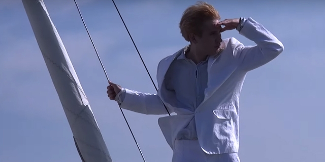 Ein junger Mann auf einer Segeljacht, in weißem Anzug, schaut in die Ferne