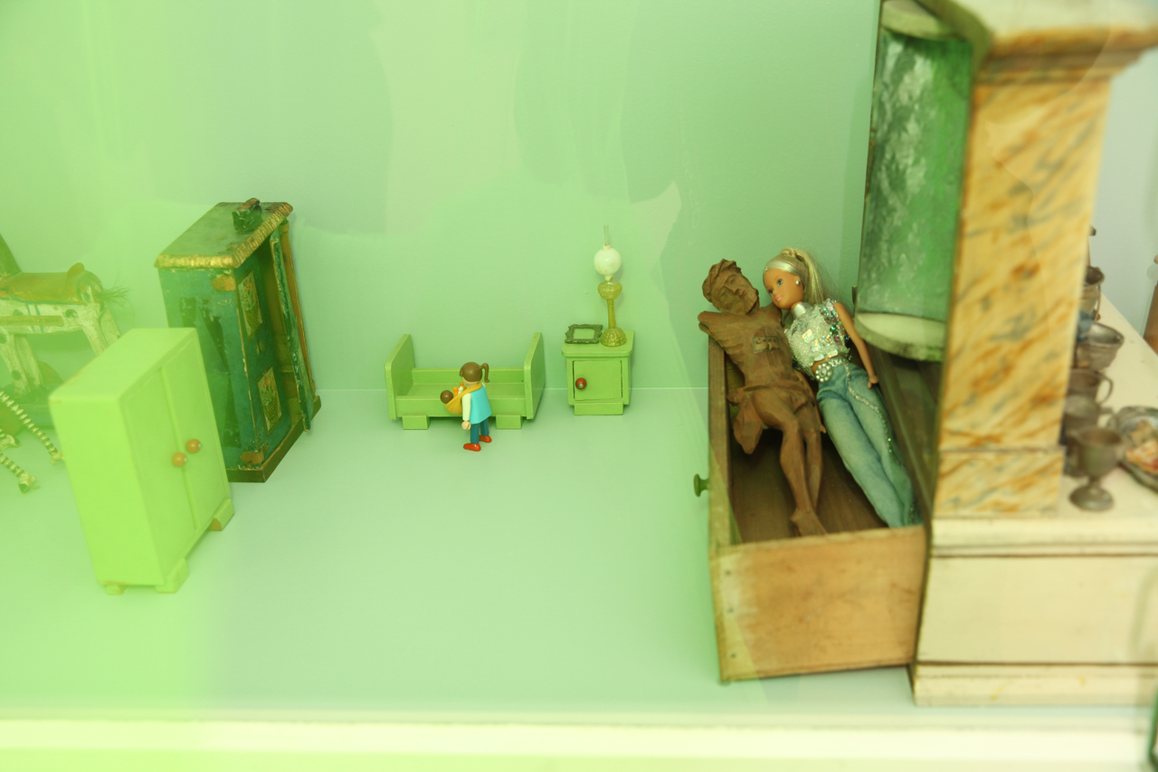 Barbie liegt neben Jesus-Figur hinter grünem Vorhang