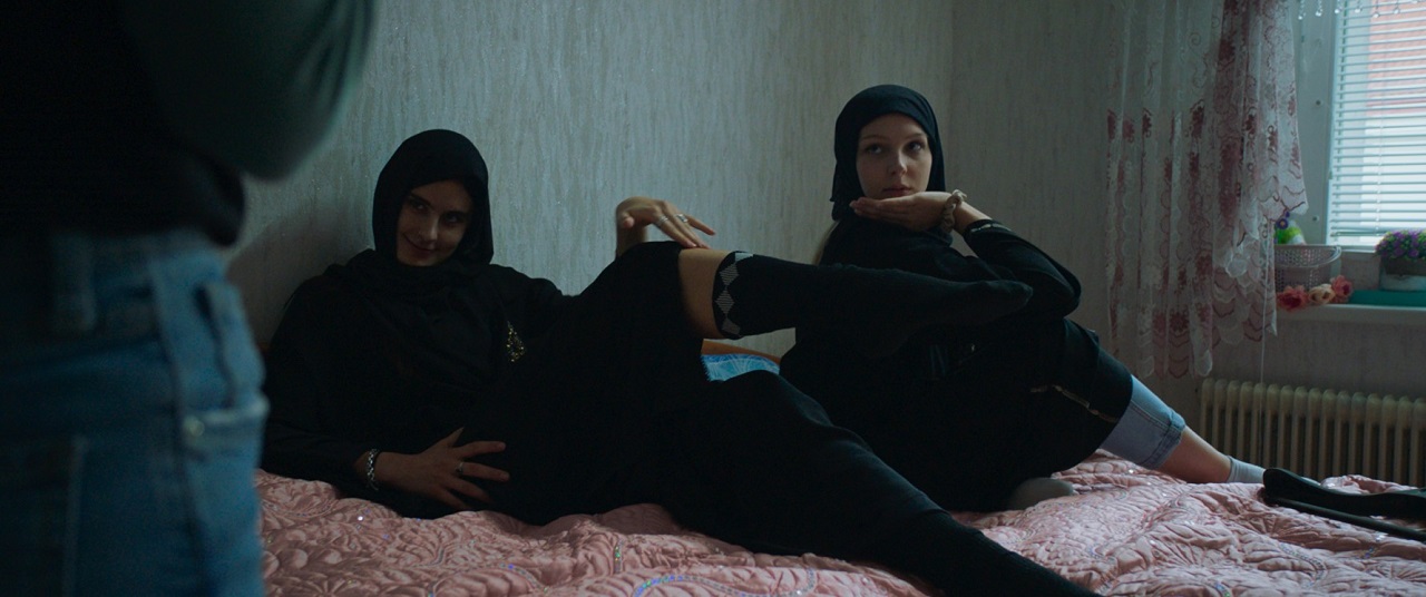Zwei Teenager mit Hijab sitzen auf einem Bett, eine präsentiert kess ein Bein und die Socke. Szene aus dem Spielfilm "Sonne" von Kurdwin Ayub.