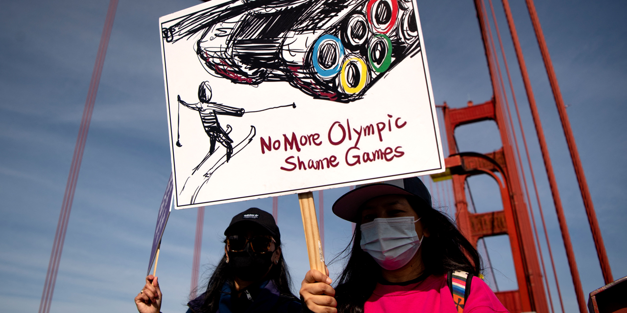 Eine Person auf einer Demo hält eine Tafel hoch. Die Tafel zeigt einen Panzer mit den olympischen Ringen und davor eine Person auf Schiern.