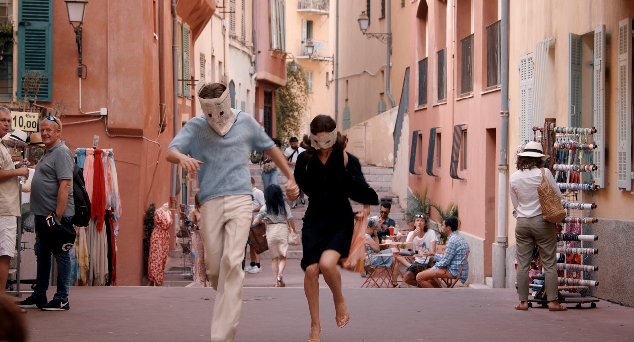 Ein junger Mann und eine Frau springen durch eine südliche Stadt, die Gesichter hinter Papiermasken versteckt. Szene aus "AEIOU - Das schnelle Alphabet der Liebe".