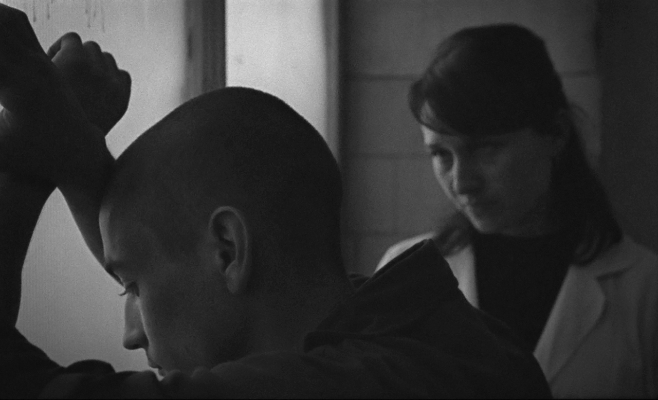 Junge Frau in weißem Kittel steht neben Mann mit geschorenem Kopf. Szene aus "Erasing Frank".