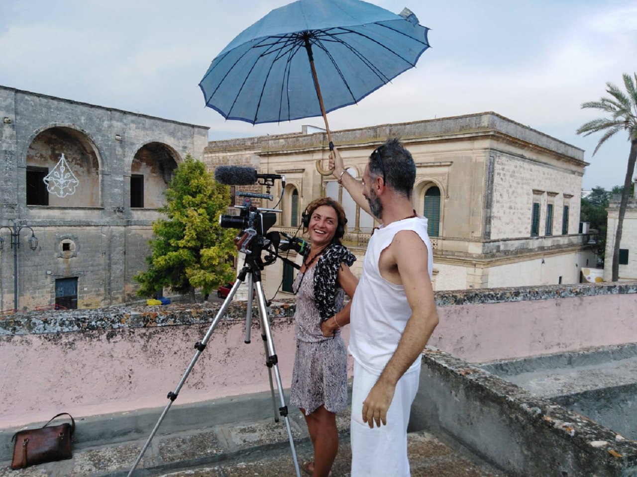 Alessandra Coppola bei Dreharbeiten für "Restanza" in Apulien, ein Mann hält ihr einen Regenschirm über die Kamera, beide lachen.