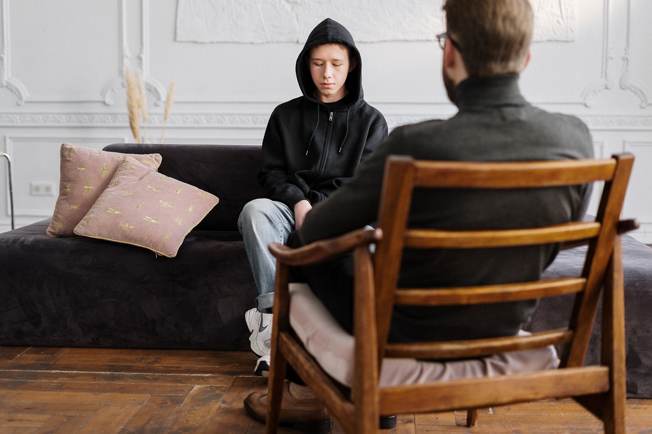 Junger Mann mit Hoodie auf Couch sitzend, Psychiater ihm gegenüber mit dem Rücken zum Betrachter