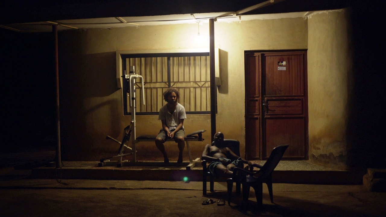 Jide Tom Akinleminu sitzt vor dem Haus seines Vaters in Nigeria, der Vater liegt etwas fern von ihm in einem Sessel. Es ist Nacht. Szene aus der Doku "When a farm goes aflame".