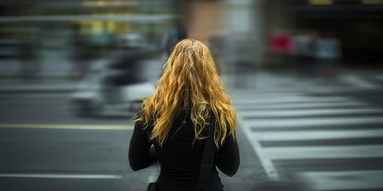 Junge Frau mit rötlichen Haaren in schwarzem Mantel von hinten, Straße vor ihr ist unscharf