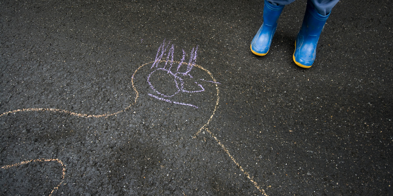 Eine Zeichnung mit Kreide auf Asphalt. Daneben ein Kind in Gummistiefeln.