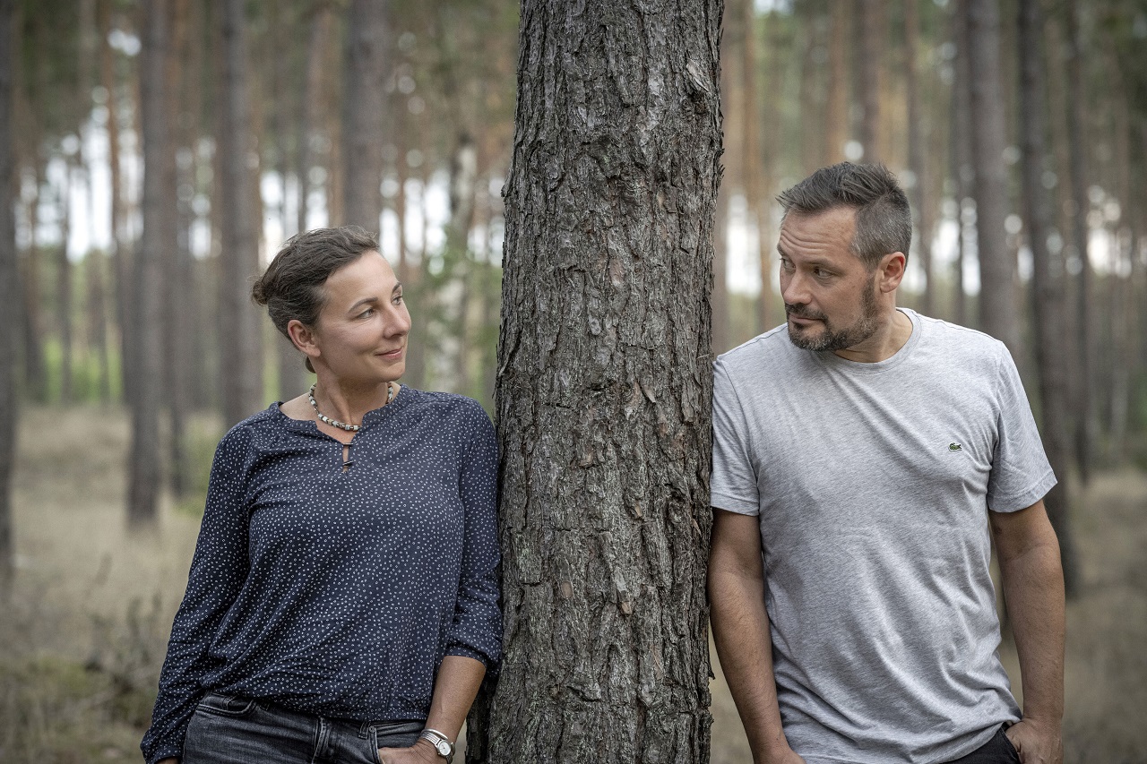 Juli Zeh und Simon Urban stehen neben einem Baum im Wald und schauen einander an.