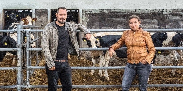 Juli Zeh und Simon Urban vor einem Stall, Kühe stehen im Freien hinter einem Zaun.