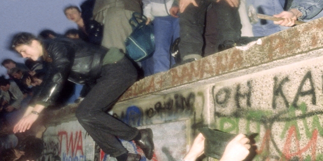 1989: Viele Menschen auf und vor der Berliner Mauer, eine Person hämmert Stücke aus der Mauer, eine andere springt herunter.