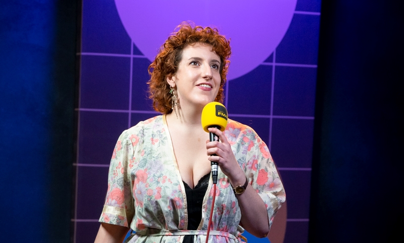 Lena Hödl auf der Standup-Bühne, sie trägt einen geblümten Kimono und hält ein gelbes FM4-Mikro in der Hand und blickt mit leichtem Lächeln nach oben