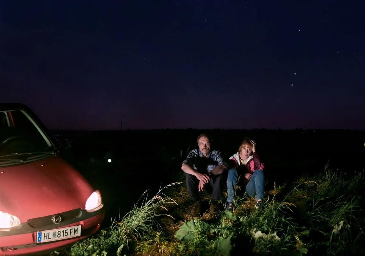 Zwei Menschen sitzen des Nachts im Gras, beleuchtet von den Scheinwerfern eines parkenden Autos